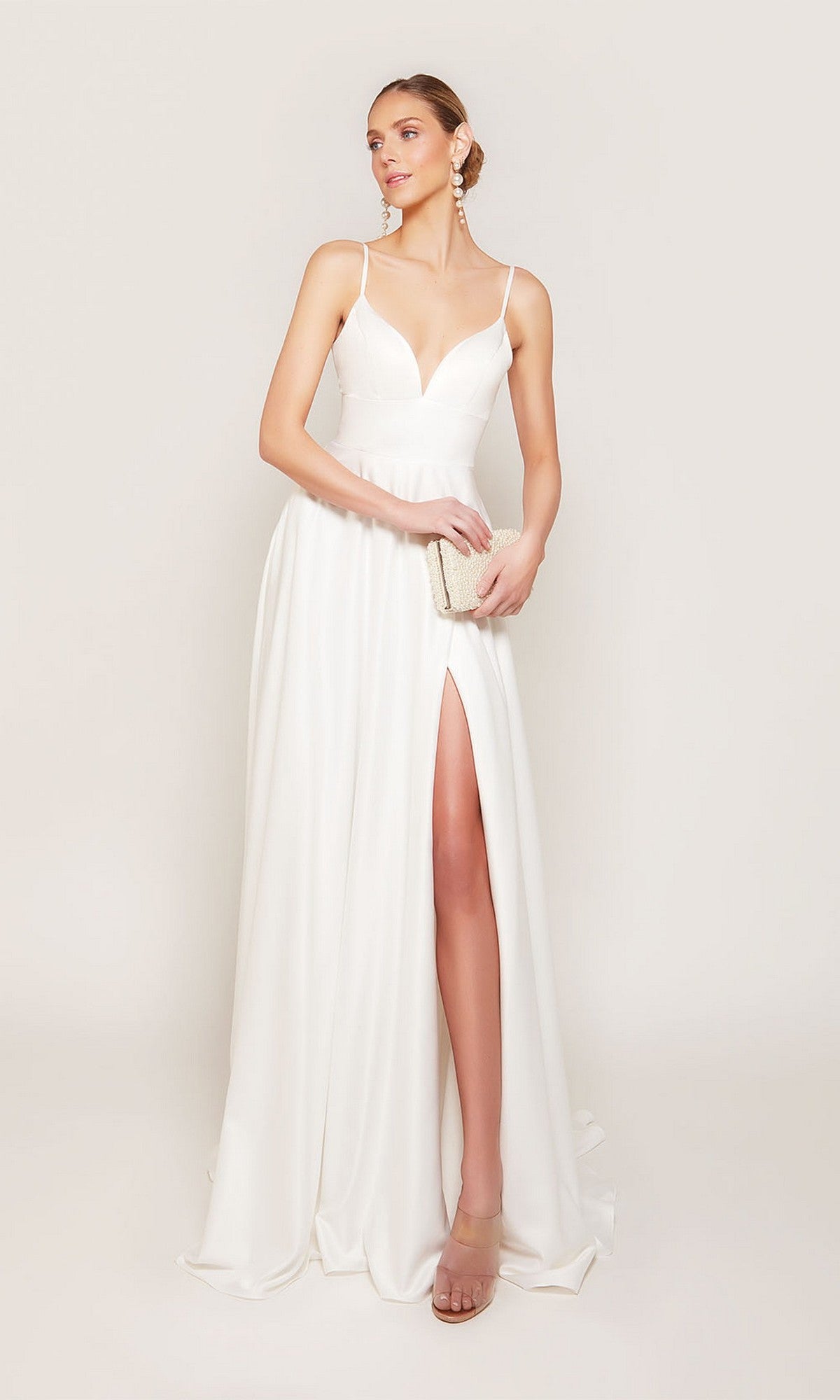 A woman stands against a plain light background wearing the Alyce 7095 long white dress featuring satin fabric, a deep neckline, and high leg slit, holding a small beige clutch.